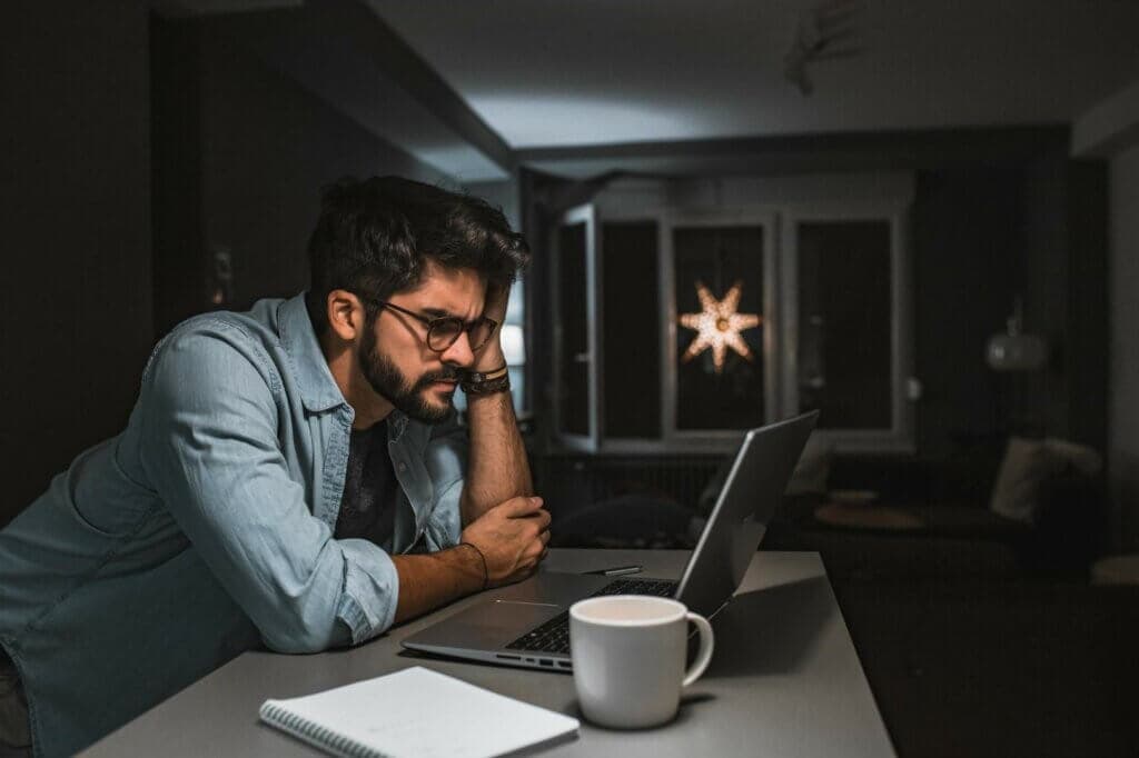 Photo: A man in a dark room at night leans on his desk and looks at his laptop with frustration.
